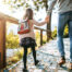 A young girl wearing a backpack holds her father's hand as they walk along a leaf-strewn stone path on a sunny fall day.
